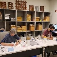 Two volunteers sit at a long table gluing plant leaves onto archival paper to be filed as reference materials in the Herbarium Library.